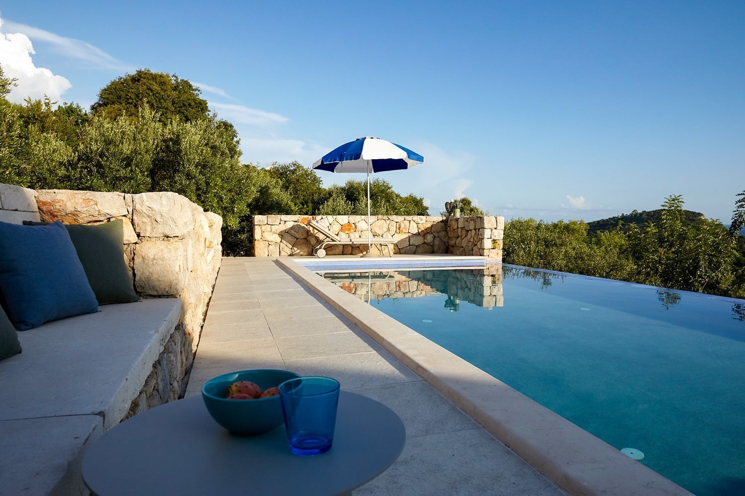 Une piscine bordée d’un muret en pierre, une chaise longue et un parasol.