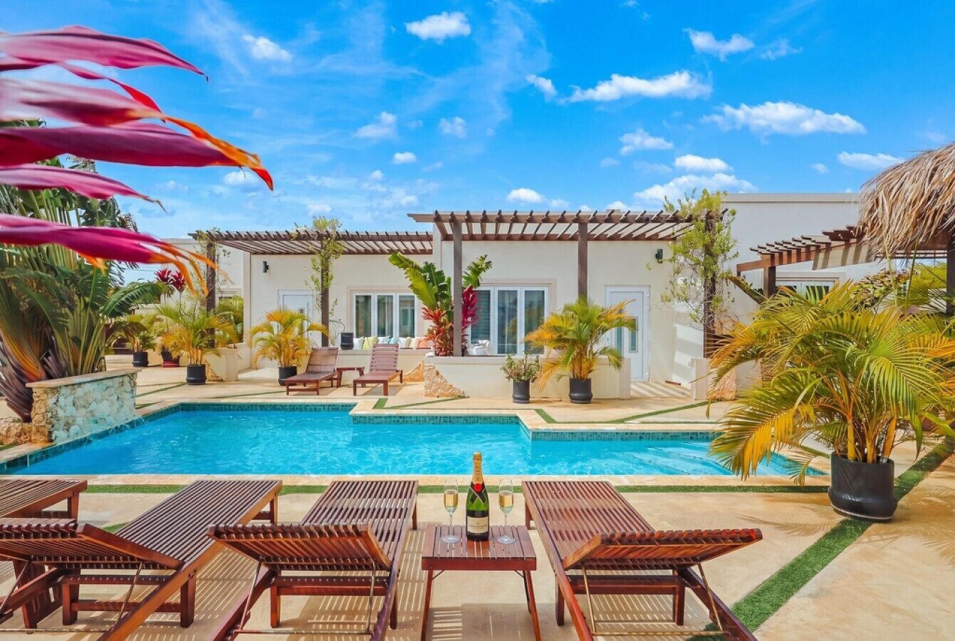 Ein Poolbereich mit Liegestühlen, einem Tisch mit Flasche und Gläsern, und einem Gebäude mit einer Pergola.