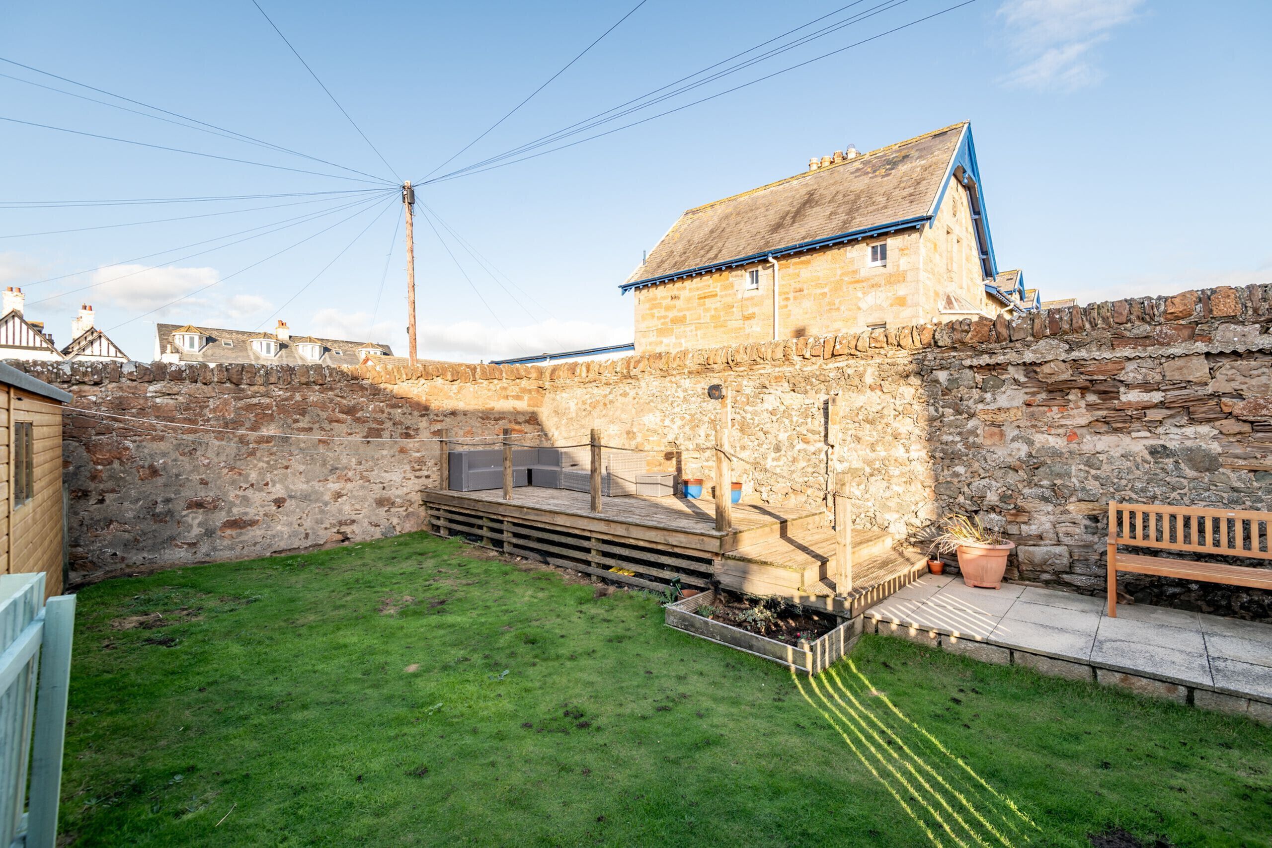A stone building with a wooden deck and a bench, surrounded by a stone wall and a grassy area.