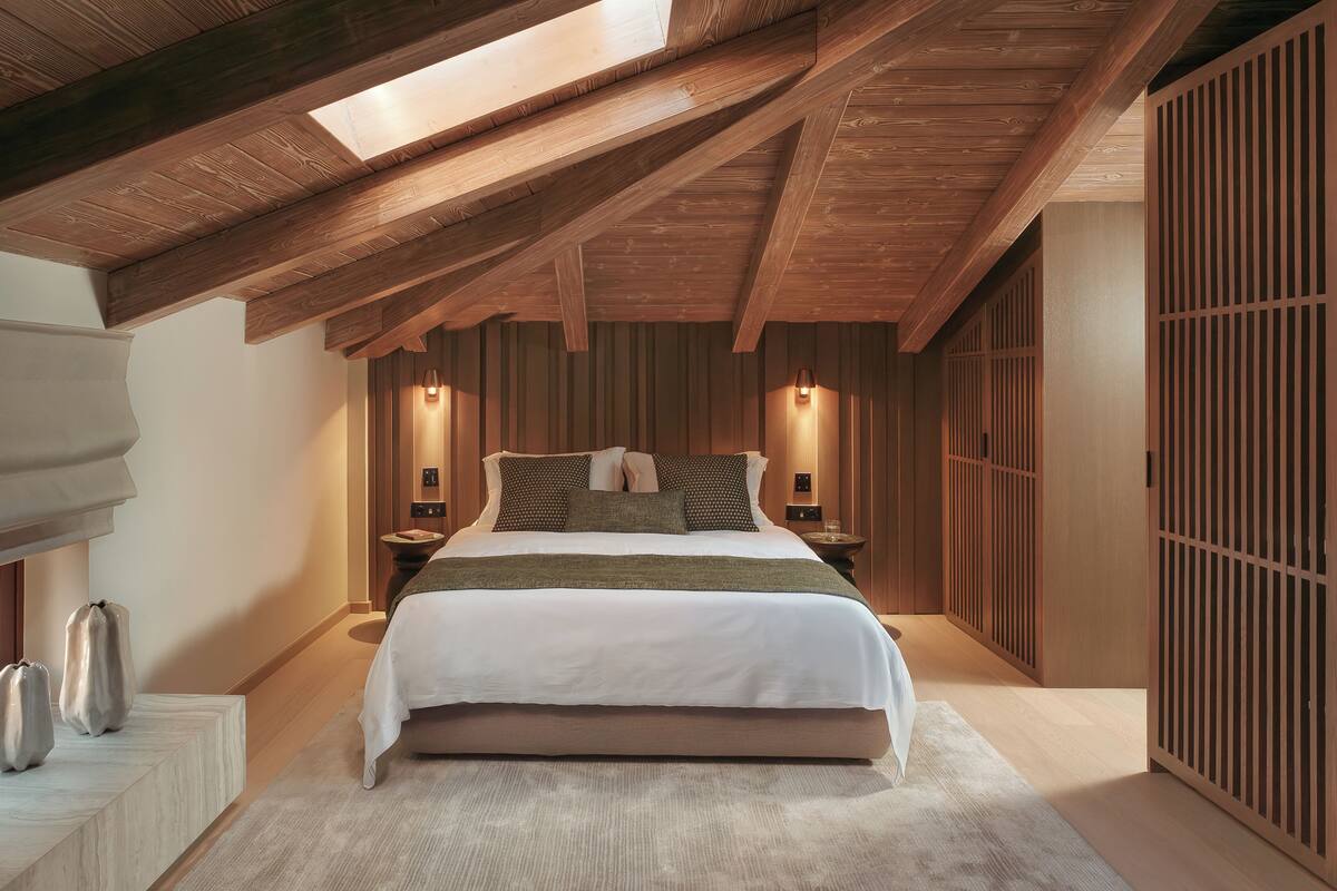A bedroom with a sloped ceiling, a bed, bedside tables, wall-mounted lights, and a large window.