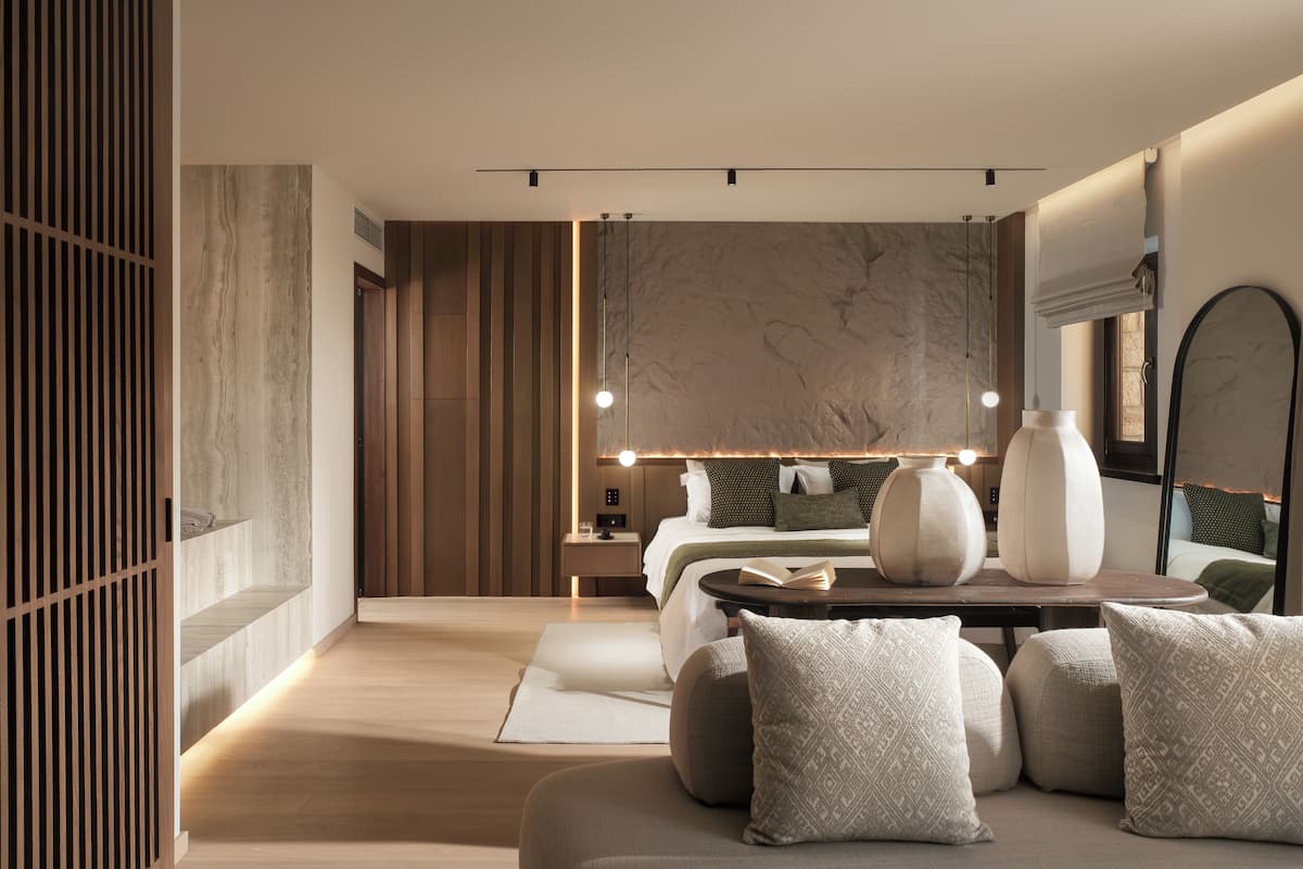 A modern hotel room with a wooden floor, a large bed with a headboard, bedside tables, and a seating area with cushions.