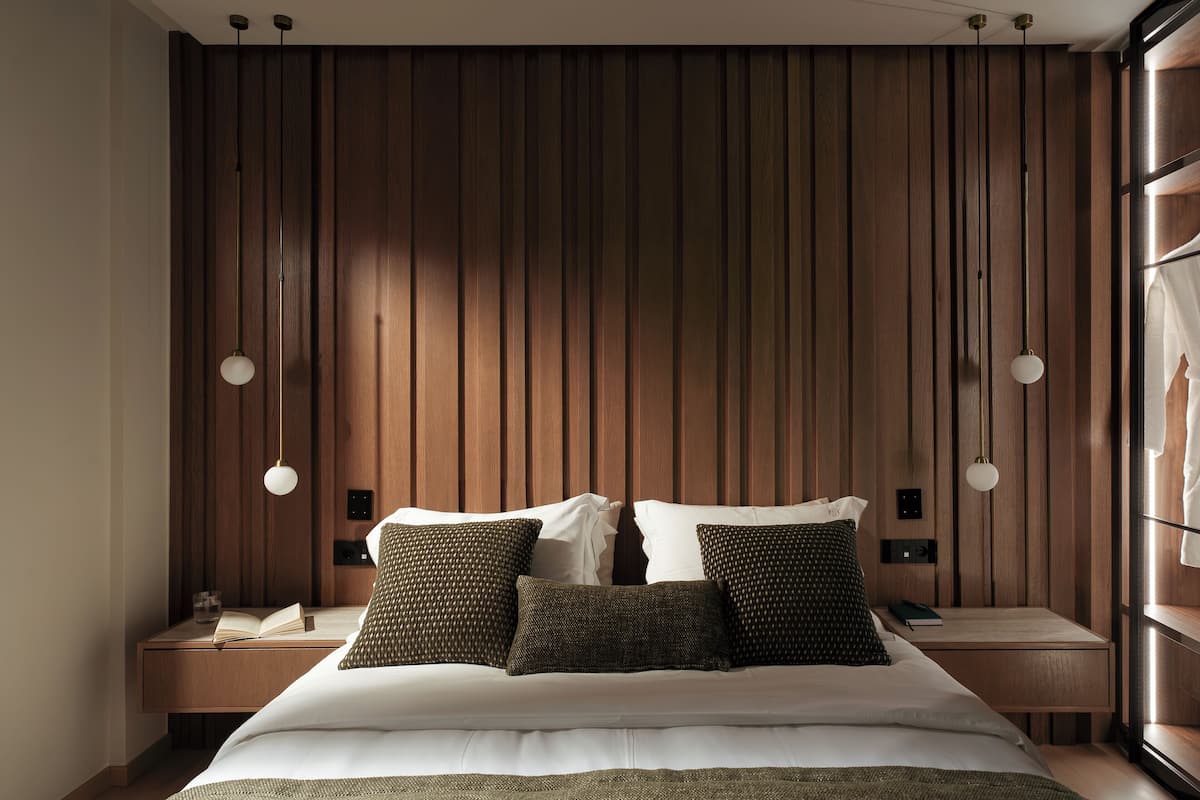 A modern bedroom with a wooden headboard, a large bed with white linens, two bedside tables, and pendant lighting.