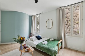 A modern bedroom with a bed, a bedside table with a vase of flowers, and a view of trees through the window. - Lavie Maison 17th district (Paris)