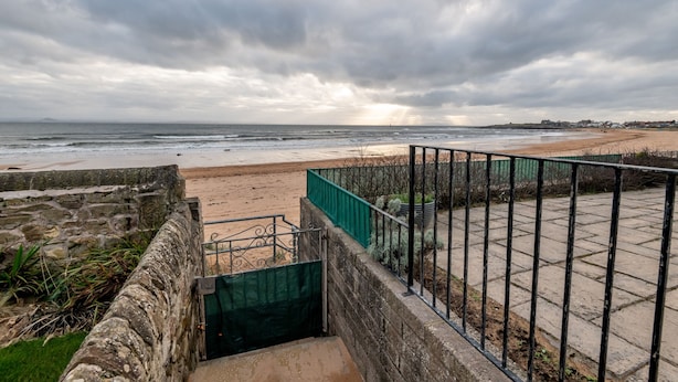 Una passeggiata costiera con muro in pietra, ringhiera metallica e vista sulla spiaggia e l'oceano sotto un cielo nuvoloso.