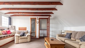 Un accogliente soggiorno con divano, poltrona e tavolino. Si vede una finestra con tende, un termosifone e travi in legno al soffitto.