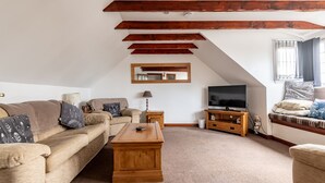 Un soggiorno con soffitto alto, travi a vista in legno, un'ampia televisione a schermo piatto, un divano, un tavolino da caffè e una finestra con tende.