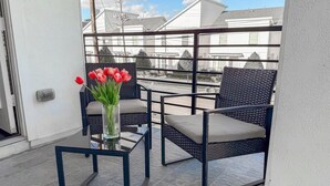 Ein Balkon mit zwei Korbstühlen, einem Glastisch, einer Vase mit roten Blumen und Blick auf Wohnhäuser.