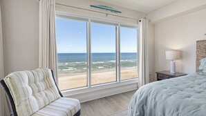 Ein Schlafzimmer mit großem Fenster mit Blick auf einen Strand, ein Bett, ein Sessel und ein Nachttisch mit Lampe.
