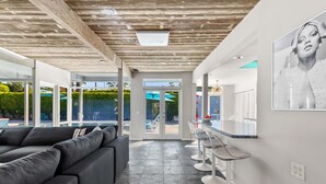 A modern interior with a sectional sofa, a bar area with stools, and a view of the outdoors. - Azure Palms at Deepwell – Luxury Pool Retreat (Palm Springs)