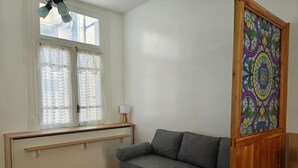 Een kamer met een grijze bank, een houten kast, een raam met dunne gordijnen en een kleurrijk glas-in-loodraam.