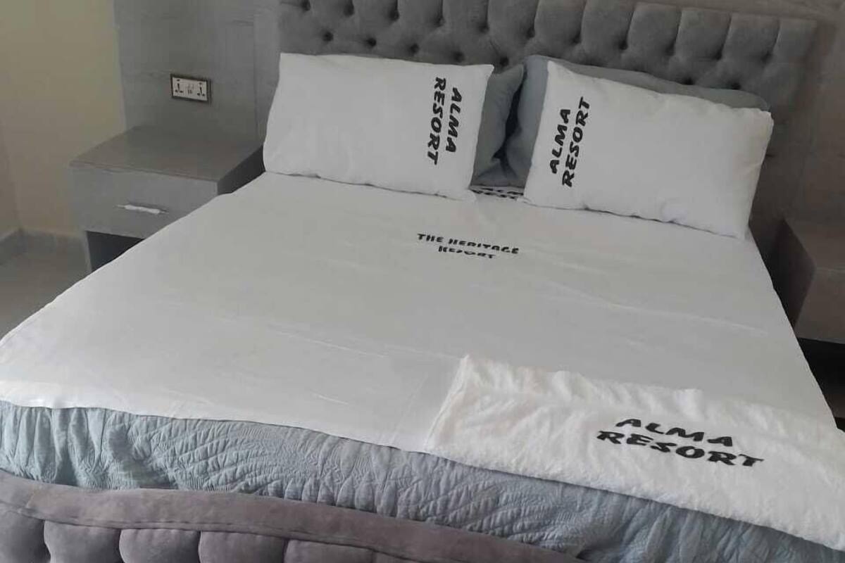 A bed with a grey tufted headboard, two pillows with 'ALMA RESORT' text, and a white bedspread.