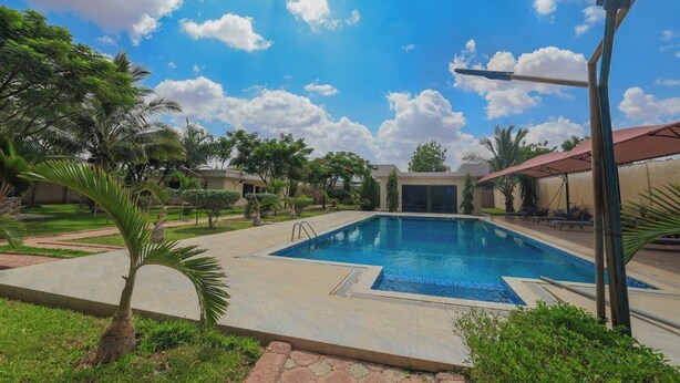 A rectangular swimming pool with a tiled deck, surrounded by palm trees and a clear blue sky with clouds. - The Heritage Resort (Wajir)