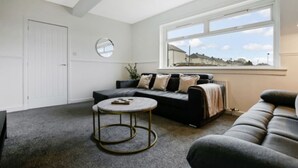 A modern living room with a sofa, a round coffee table, and a large window with a view. - Free Parking | Central | Sleeps 6 | 3 Bedrooms (North Lanarkshire)