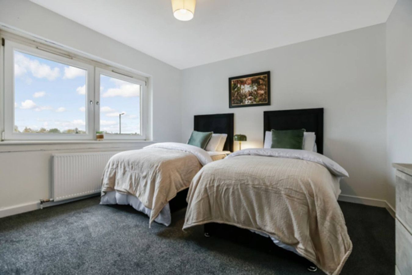 A bedroom with two beds, a radiator, a window with a view, and a framed picture on the wall.