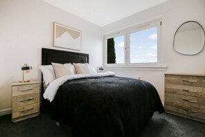 Un dormitorio con cama, mesita de noche, cómoda, ventana con vistas y un cuadro en la pared.