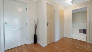 A hallway with wooden flooring, white walls, and a bathroom visible through an open door.