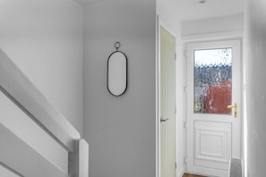 Un pasillo con una puerta blanca, un objeto ovalado pequeño en la pared y una ventana que da a vista de árboles.