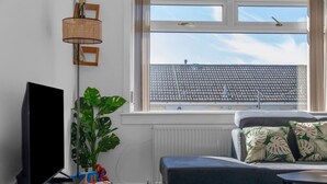 A living room with a flat-screen TV, a potted plant, a sofa with patterned cushions, and a window with a view of a tiled roof.