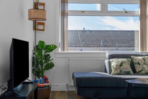 A living room with a flat-screen TV, a potted plant, a sofa with patterned cushions, and a window with a view of a tiled roof.