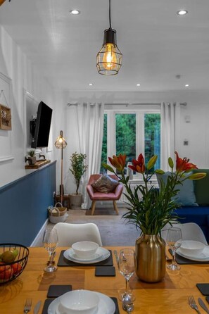 A dining area with a wooden table set for two, a vase of flowers, and a television on the wall.