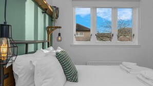 A bedroom with a bed, pillows, a bedside lamp, a window with a view of a house, and a radiator. - Free Parking | Central | Sleeps 5 | 2 Bedrooms (West Lothian)
