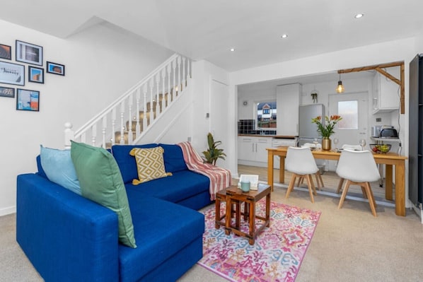 A modern living room with a blue sofa, a dining area with a wooden table and chairs, and a kitchen in the background.