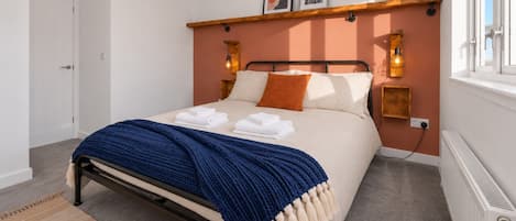 Un dormitorio con una cama, una colcha azul, un almohadón blanco, una estantería de madera con libros y un aplique de pared.