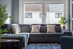 Un salon moderne avec un canapé gris, des coussins à motifs, un tapis bleu et des plantes en pot.