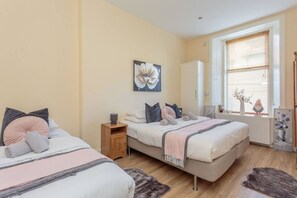 Une chambre avec deux lits, une table de chevet, un radiateur, une fenêtre avec des stores et un vase à fleurs.