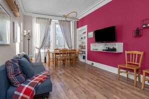 A living room with a sofa, dining table, chairs, a TV mounted on a pink wall, and a bookshelf. - Free Parking | City Centre | Sleeps 9 | 3 Bedrooms (Edinburgh)
