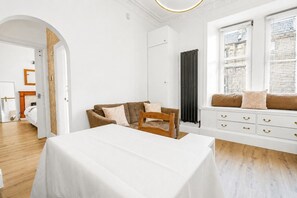 A hotel room with a white table, a brown sofa, a wooden chair, a white dresser with gold handles, and a radiator. - Stunning 1-Bedroom Flat in Edinburgh | Sleeps 3 (Edinburgh)
