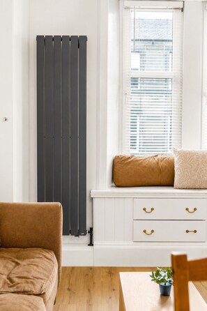 A room with a radiator, a window with blinds, a cushioned bench, and a wooden table with a plant. - Stunning 1-Bedroom Flat in Edinburgh | Sleeps 3 (Edinburgh)