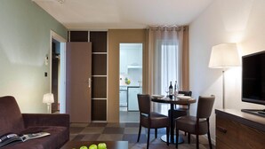 A hotel room with a sofa, a dining table with chairs, a TV, and a kitchen area visible through an open door. - Elegant Apartment Near Geneva (Ferney-Voltaire)