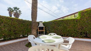 Eine Terrasse mit einem runden Tisch und weißen Stühlen, umgeben von einem Holzboden und einem Heckenstreifen.