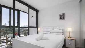 Ein modernes Hotelzimmer mit einem großen Bett, zwei Nachttischen mit Lampen, Blick auf die Stadt durch raumhohe Fenster und einem kleinen gerahmten Bild an der Wand.