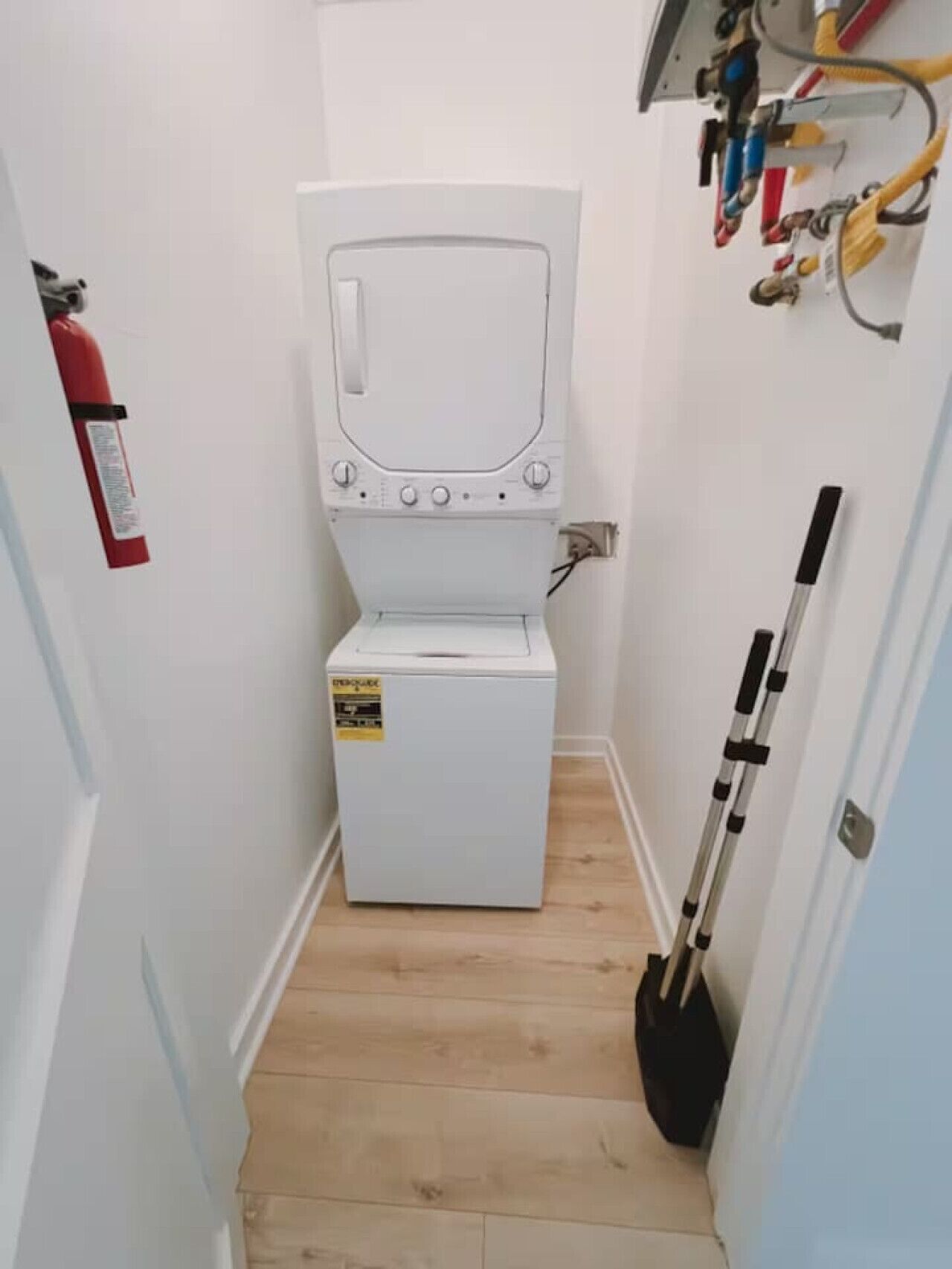 A compact laundry room with a white washing machine, a fire extinguisher, and a broom leaning against the wall.