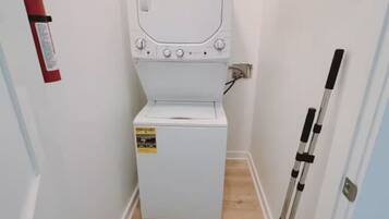 A compact laundry room with a white washing machine, a fire extinguisher, and a broom leaning against the wall.