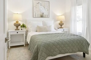 A bedroom with a bed, two bedside tables, a nightstand, a window with curtains, and a wall-mounted painting. - Weekend Escape on the Coast - Relax & Unwind (Pass Christian)