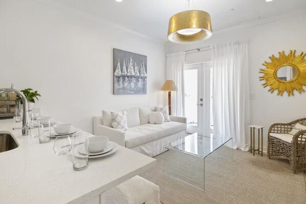 A modern, minimalist living room with a white sofa, a glass coffee table, and a dining area with a white table and chairs. - Weekend Escape on the Coast - Relax & Unwind (Pass Christian)
