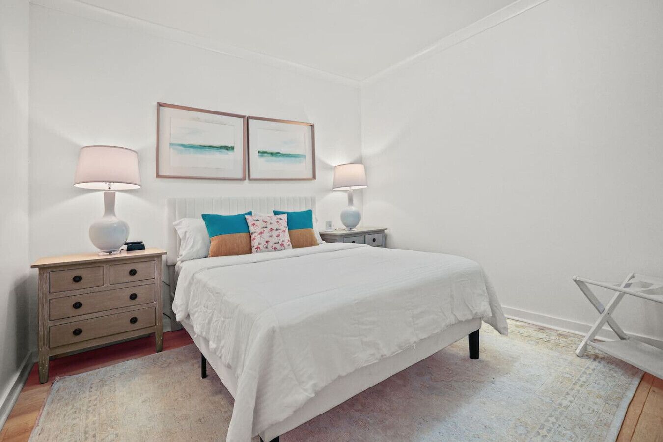 A bedroom with a large bed, two bedside tables with lamps, a small folding chair, and two framed pictures on the wall.