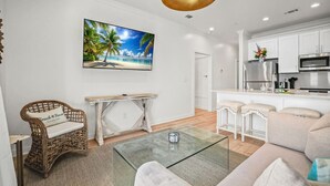 Un salon moderne avec une table basse en verre, des meubles en osier, un tableau représentant une scène de plage et un coin cuisine doté de meubles blancs et d’appareils électroménagers en acier inoxydable.
