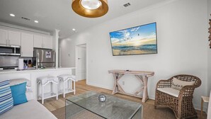 Una cucina moderna con mobili bianchi ed elettrodomestici in acciaio inossidabile, un tavolino da caffè in vetro, una sedia in vimini e un quadro raffigurante una spiaggia.