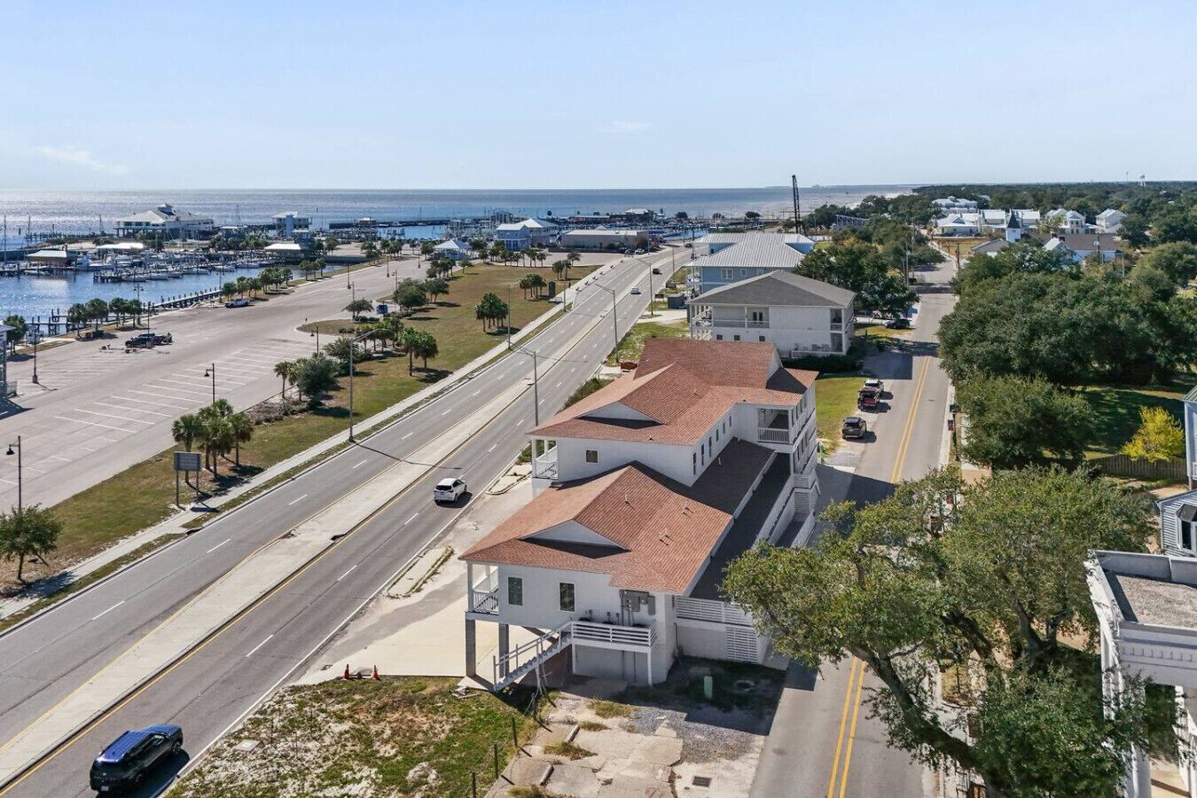 A coastal road with a marina, a white building with a red roof, and residential houses in the background.