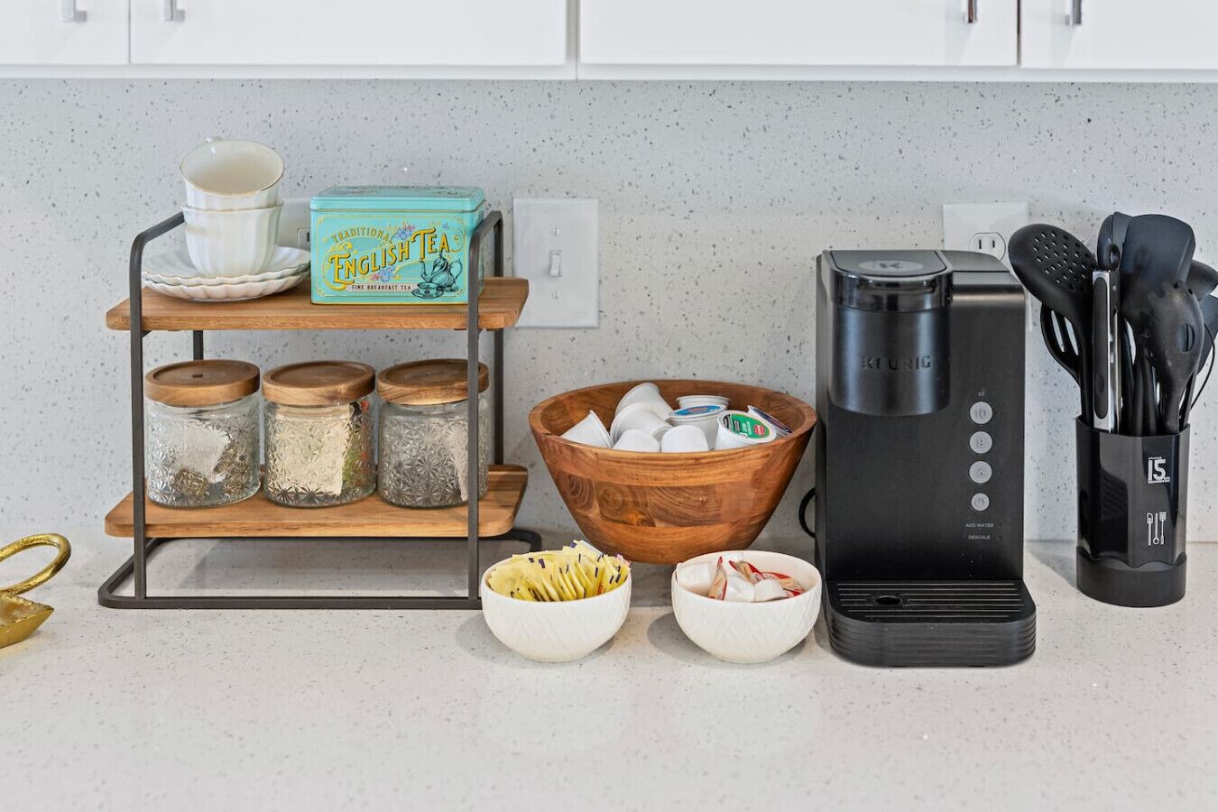 A kitchen counter with a coffee maker, a tea box, jars, a wooden bowl, and a utensil holder.