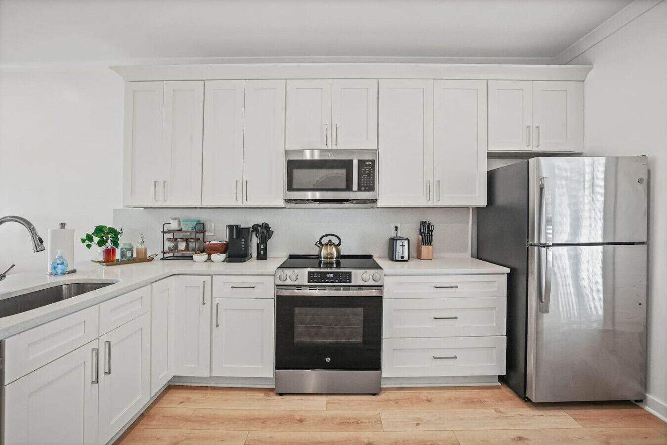A modern kitchen with white cabinets, a stainless steel refrigerator, a microwave, a stove, and a sink.