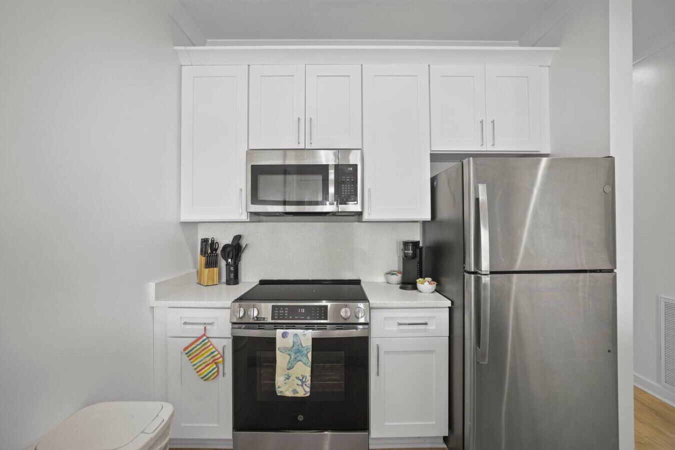 A compact kitchen with white cabinets, a microwave, a stainless steel refrigerator, an oven, and a coffee maker.
