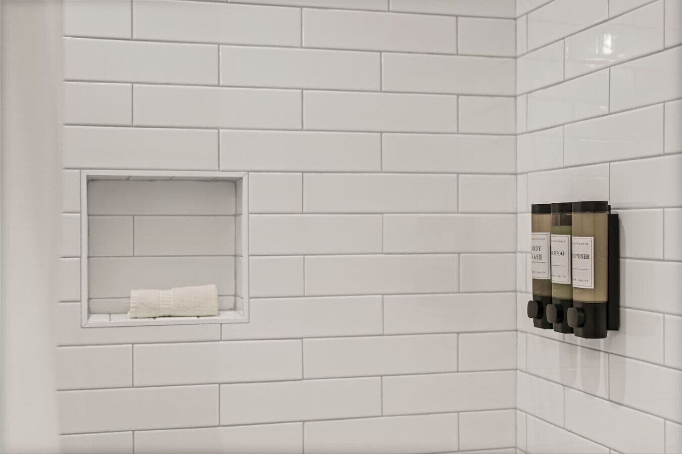 Een badkamer met witte metro tegels, een klein raam met een handdoek, en een wandbevestigde zeepdispenser met flessen shampoo en zeep.