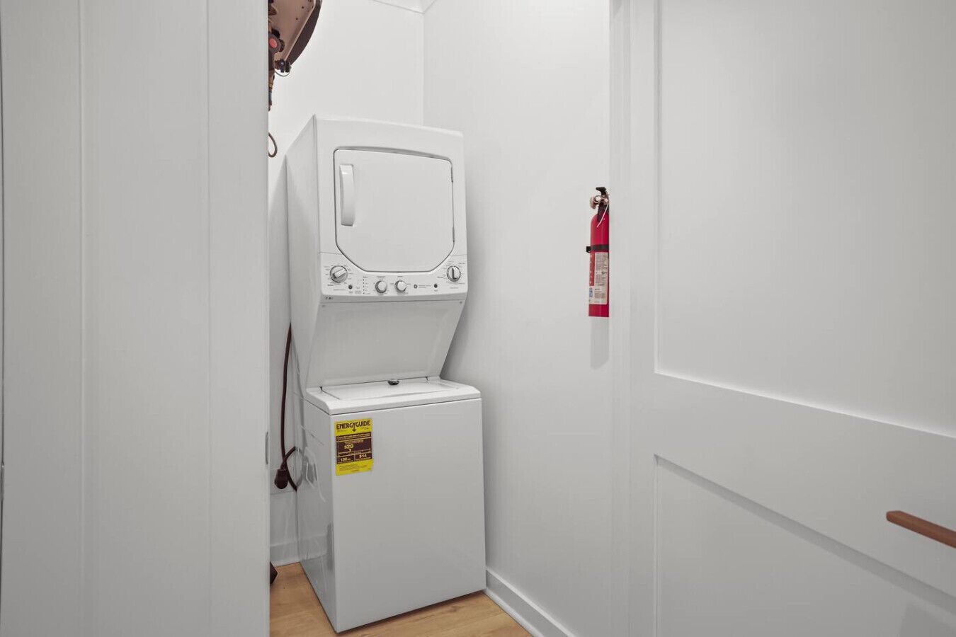 A compact, white laundry room with a front-loading washer and dryer, a fire extinguisher on the wall, and a wooden floor.