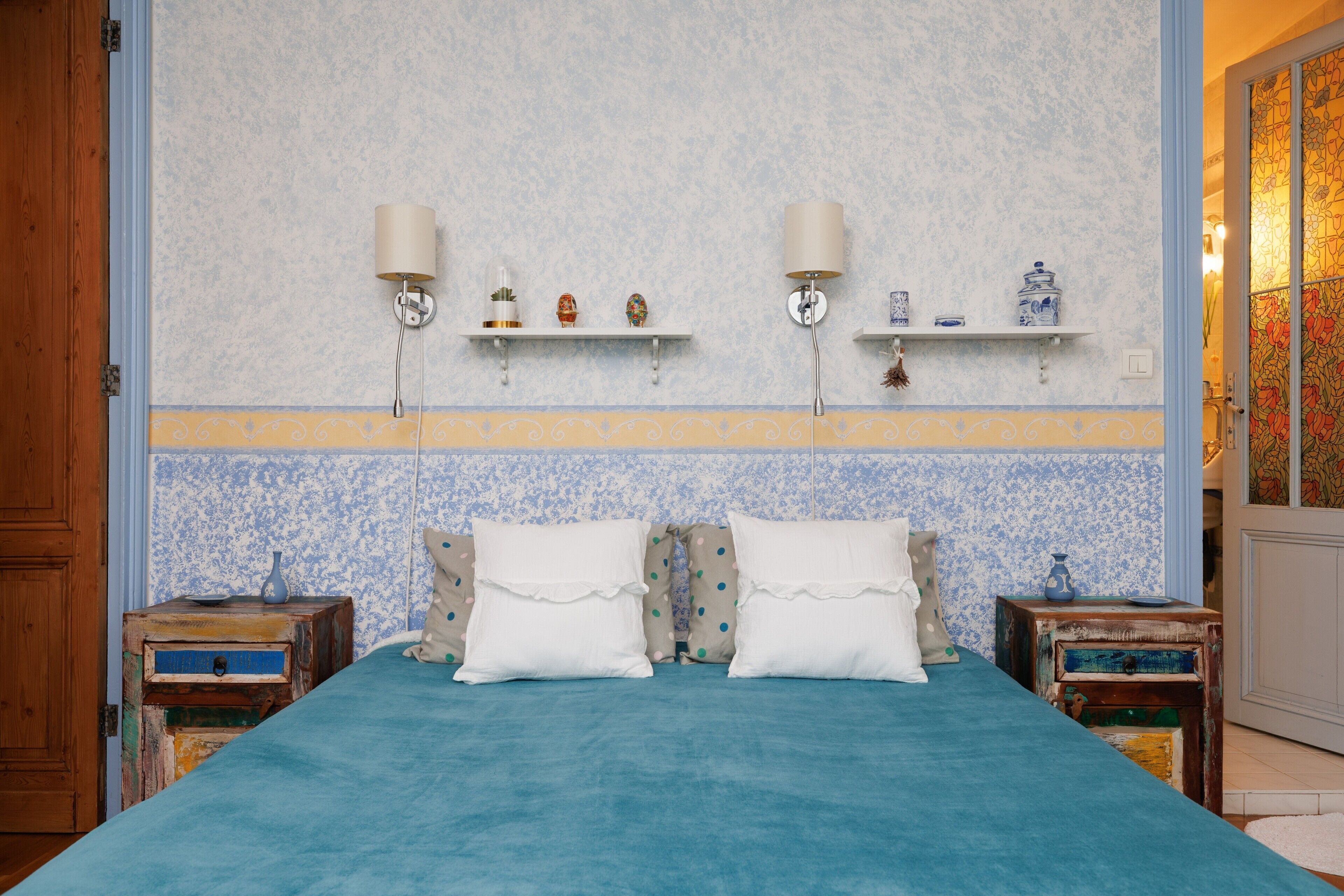 Un dormitorio con una cama, dos mesitas de noche, estantes en la pared y un papel tapiz decorado.