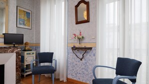 Une pièce comprenant une télévision, deux fauteuils bleus, une petite table avec un vase de fleurs, un miroir et un élément décoratif mural.